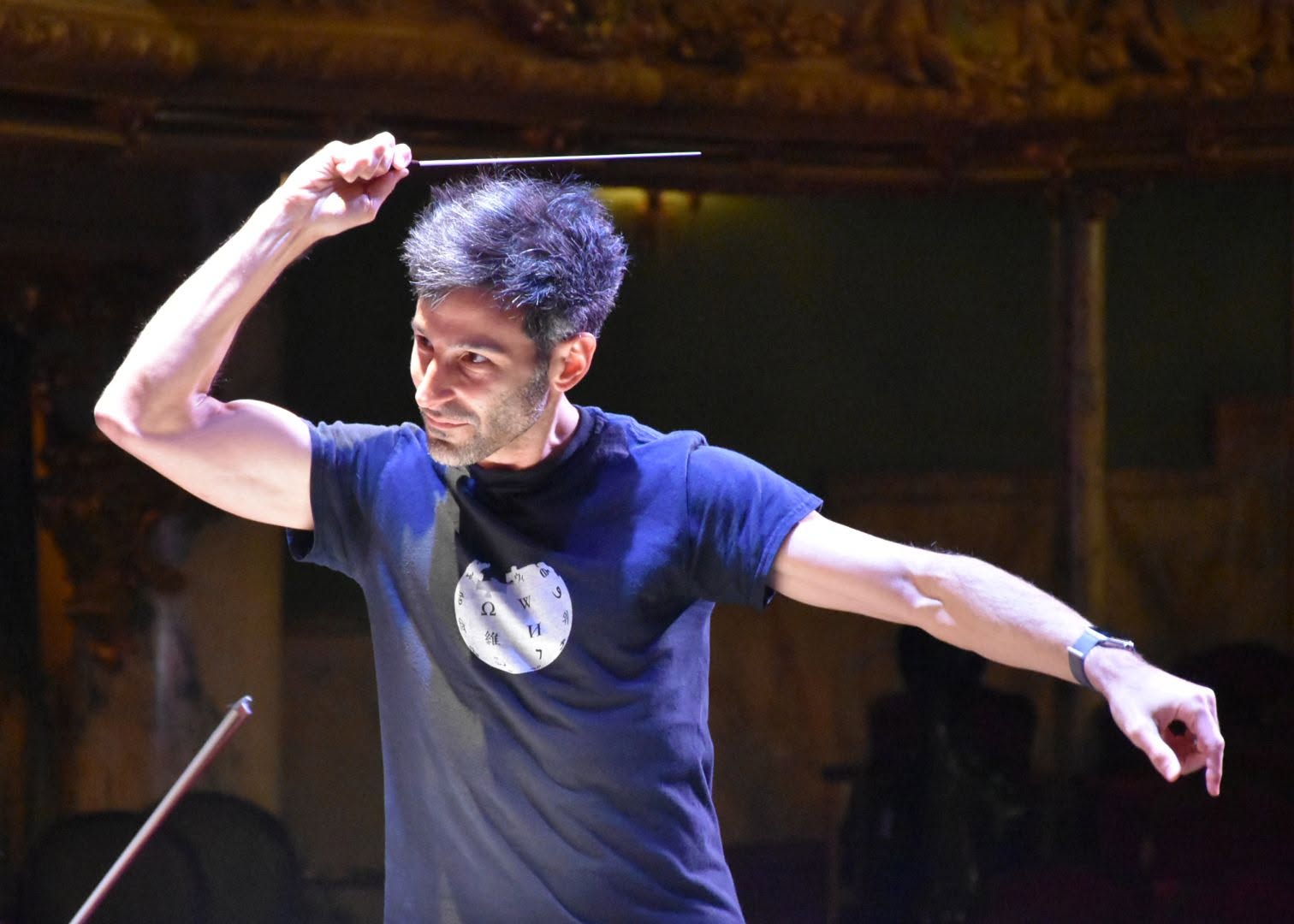 Isaac Selya conducting in rehearsal, baton raised overhead and left arm extended